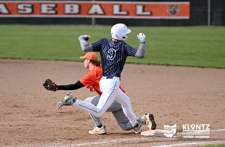 Baseball action: a baserunner in a navy striped uniform and white helmet slides into a base while a fielder in orange reaches with a glove on the dirt infield.