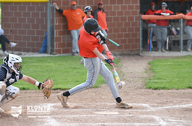 Baseball batter in an orange shirt swings at a pitch as catcher crouches behind home plate with a glove ready to tag.