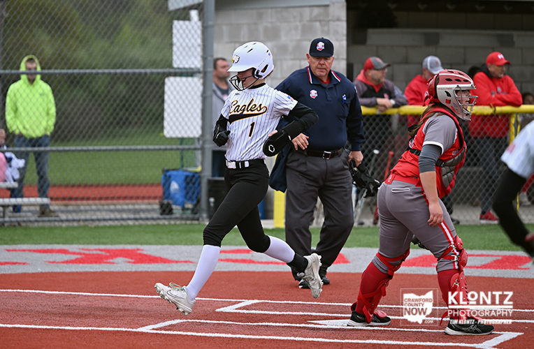 Youth softball: a runner in a white 'Eagles' jersey (#1) sprints toward home plate as the catcher in red gear and an umpire watch from the baseline behind the infield fence.
