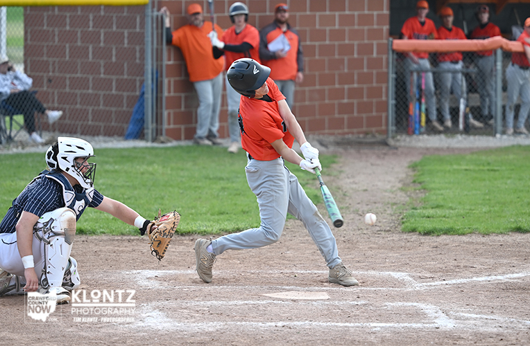 Young baseball batter in an orange shirt swings at a pitch as the catcher crouches behind home plate in protective gear.