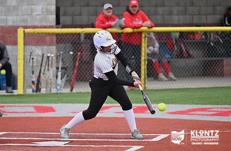 Female softball batter in white pinstripe uniform swings at a pitch on a red infield as spectators watch behind a yellow fence.