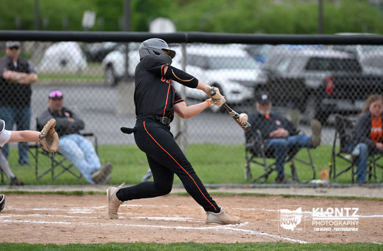 Baseball batter in a black uniform swings at a pitched ball during a game, with a catcher and spectators behind a chain-link fence in the background.