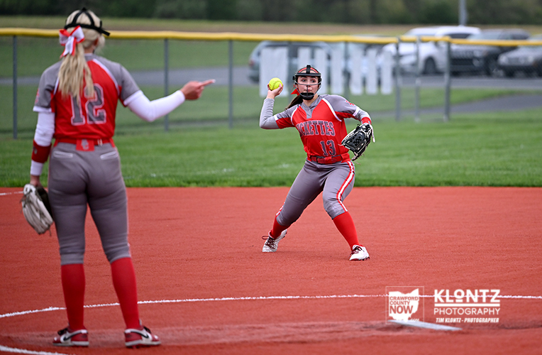 Young female softball pitcher (number 13) throws a pitch as a teammate (number 12) watches from the infield on a red dirt field.