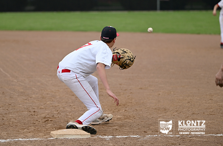 Baseball player in a white uniform crouches at first base with glove ready to field a ground ball.