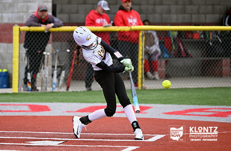 Female softball player in a white helmet and white-and-black uniform swings a bat at a pitched ball on a red dirt field, with spectators behind a chain-link fence.