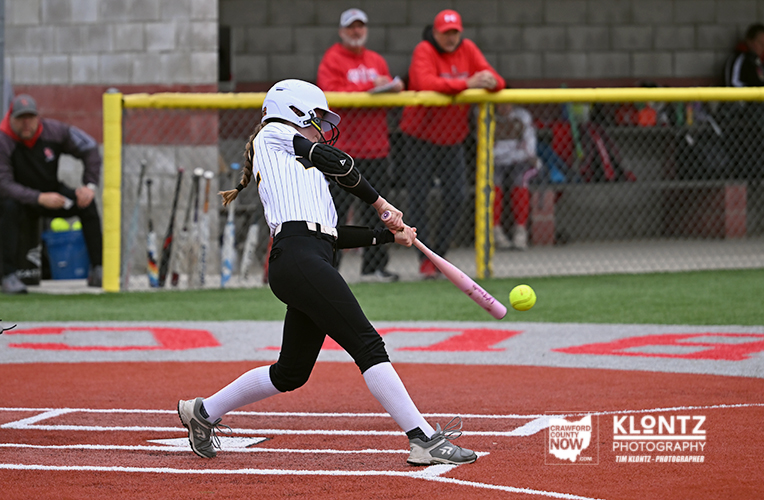 Softball batter in a white helmet and striped jersey swings a pink bat as a ball approaches the plate on a red-dirt field.