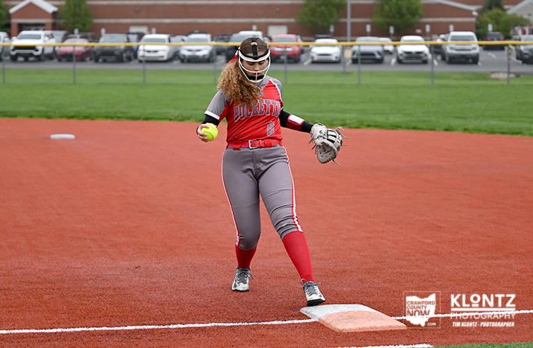Female softball player in a red uniform on first base, holding a ball in her left hand and a glove in her right, ready to throw during a game on red dirt field.