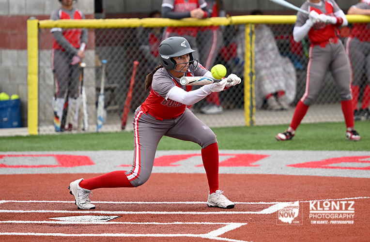 Female softball player in gray and red uniform swings at a pitch, bat making contact with a bright yellow ball near home plate.
