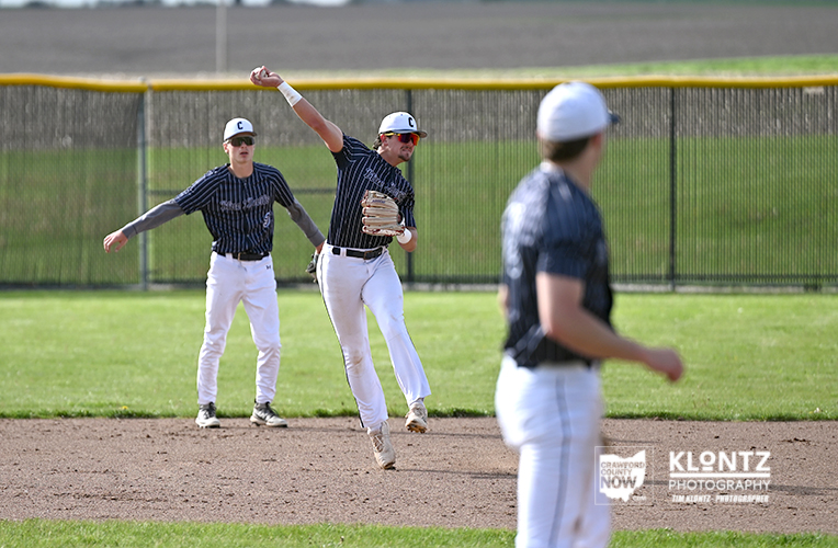 Baseball pitcher in pinstripe uniform mid-throw on the dirt infield, with teammates and a blurred fielder in the foreground.