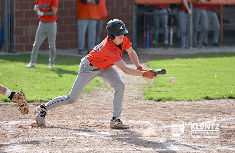Baseball batter in an orange jersey swings at a pitched ball near home plate as dust rises from the dirt.