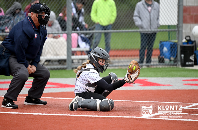 Softball catcher in full protective gear kneels at home plate, glove reaching for a rolling ball as an umpire watches nearby behind a chain-link fence