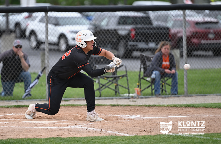 Female softball player in a black uniform and helmet is mid-swing at home plate during a game, with a chain-link fence and spectators in the background.