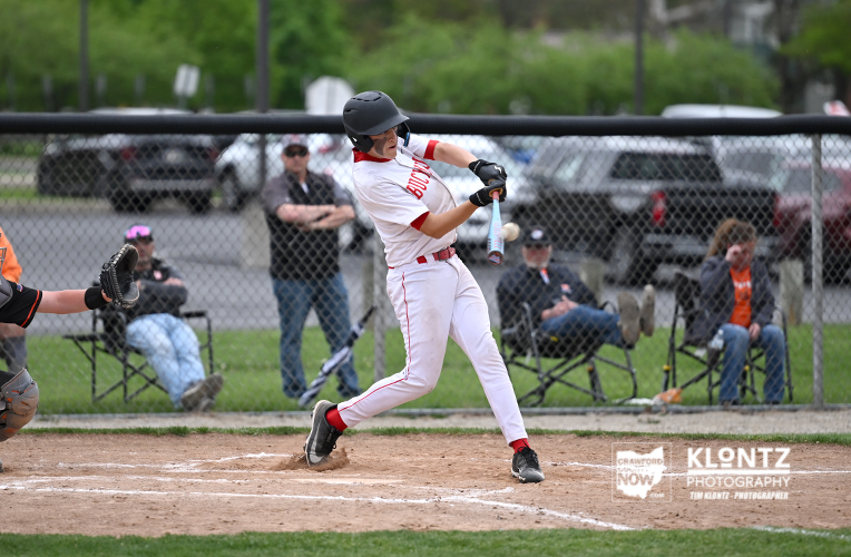 Baseball batter in white and red, mid-swing at home plate with catcher and spectators in the background.