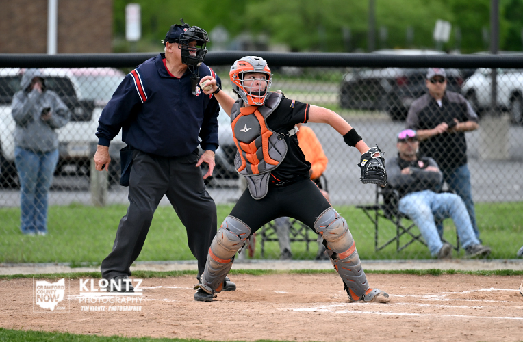 Baseball catcher in orange/gray protective gear near home plate with umpire beside him and spectators in the background behind a chain-link fence.