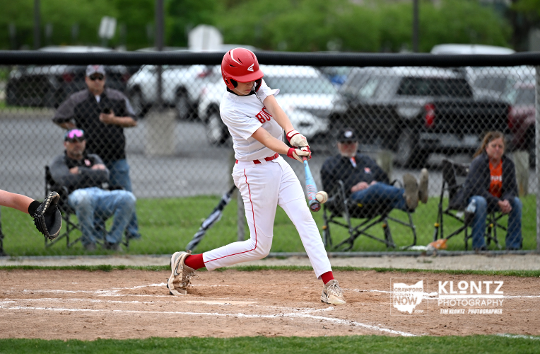 Baseball batter in white and red uniform swings at a pitch at home plate with spectators watching behind a chain-link fence