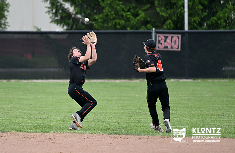Two young baseball players in black uniforms chase a pop-up ball on the infield; one leaps with glove raised while the other watches nearby on the grass and dirt field.