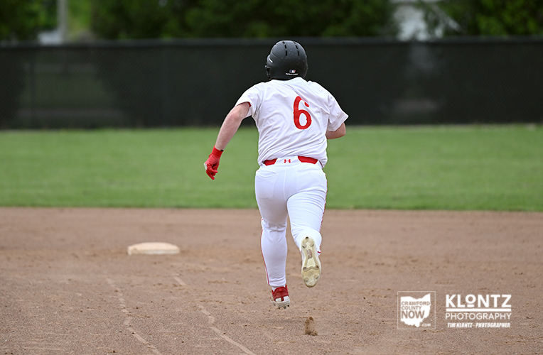 Baseball player in a white uniform with red number 6 runs toward first base on a dirt infield, seen from behind with a black helmet and red gloves.
