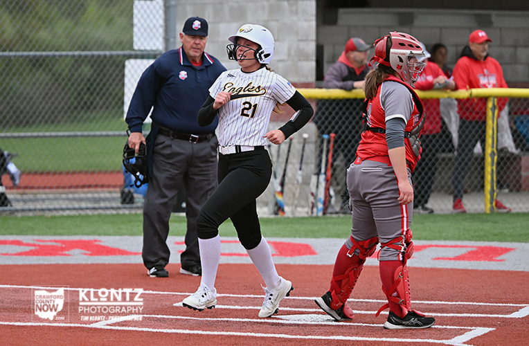 Softball scene: a runner in a white pinstripe Eagles jersey (no. 21) crosses home plate as the catcher in red gear watches, with an umpire and spectators in the background.