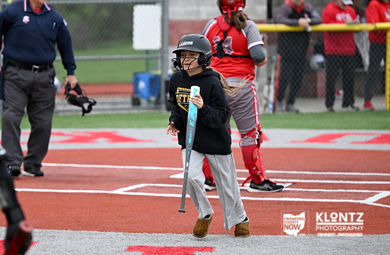 Young girl in a batting helmet and black sweatshirt holds a bat, about to swing on a youth baseball field with players and a catcher nearby in the background.