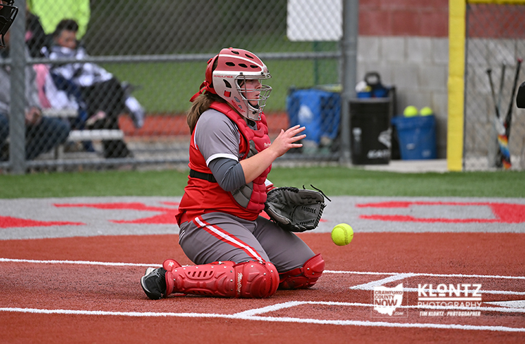 Female softball catcher in red gear kneels on the infield, ready to catch a pitch near home plate