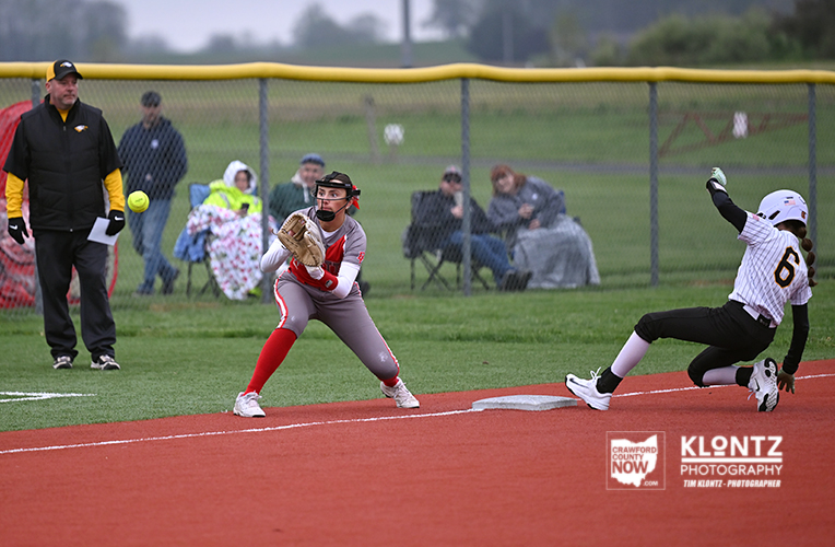 Softball catcher in gray and red crouches to field a ball near a base as a runner slides in; spectators watch from behind a chain-link fence.