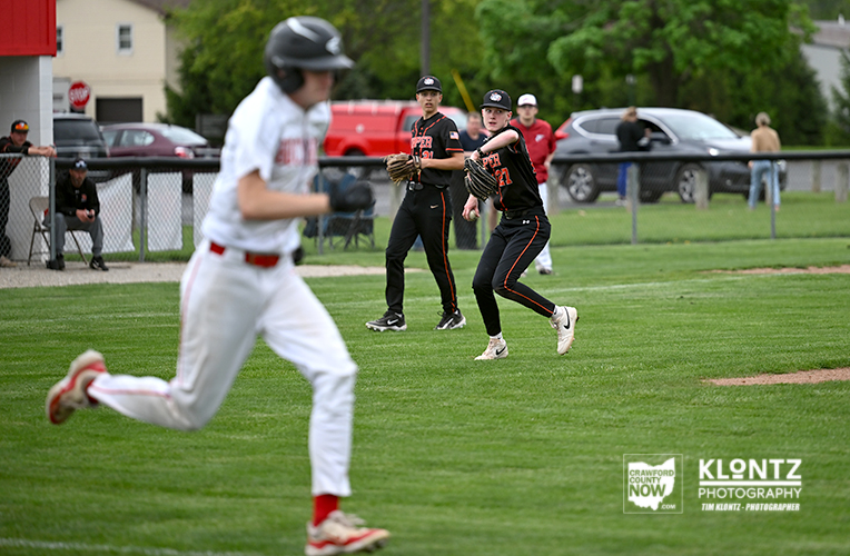 Baseball game in progress: pitcher in white uniform on the mound, with two black-uniform fielders ready at the infield as spectators watch behind a fence.