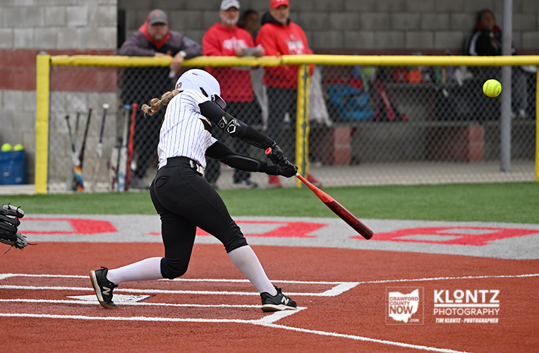 Female softball player in white pinstripes swinging a red bat at a pitch on a dirt field.
