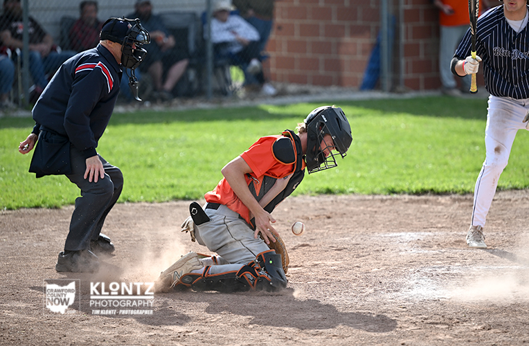 Baseball catcher in orange gear kneels in dirt as a pitch slips by, with an umpire behind and a batter waiting at the plate nearby.