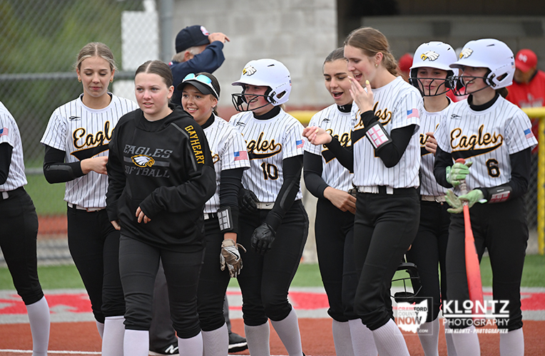 Girls softball team in white jerseys huddle on the field after a game, smiling and talking.