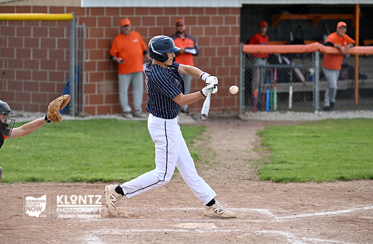 Baseball batter in a navy pinstripe uniform swings at a pitch at home plate, with the catcher’s mitt visible to the left and players watching from the dugout behind.
