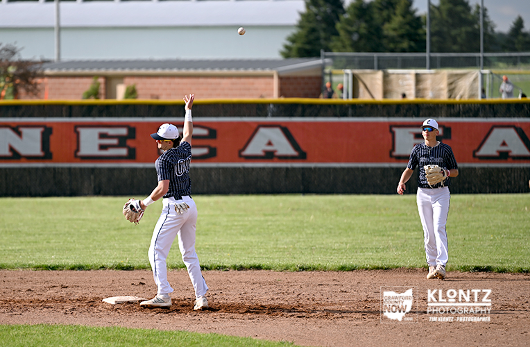 Two baseball players on the infield: one throws with their arm raised and ball in the air while the other stands ready at the grass and dirt field, with a red wall in the background.