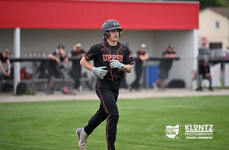 Baseball player in a black uniform with 'UPPER' runs on the grass with a helmet and glove during a game; dugout and spectators in the background.