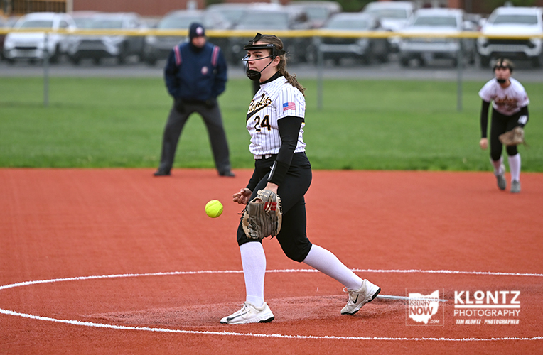 Female softball pitcher on the mound in a striped uniform, glove readied, about to throw a pitch as a yellow ball sails midair