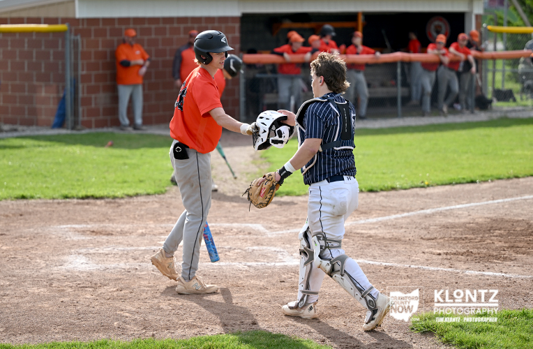 Baseball players share a high-five near home plate as the runner in red helmet meets the catcher in dark gear, with teammates in the dugout in the background.