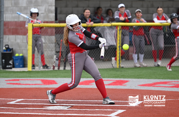 Female softball player in gray and red uniform swings at a pitch, ball in midair near the bat on a dirt infield with spectators behind a chain-link fence.