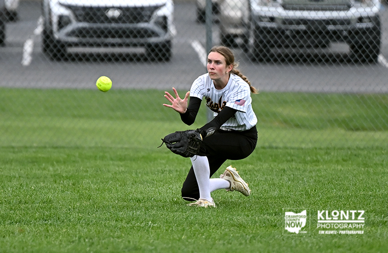 Female softball player in a white uniform crouches with glove ready, watching a yellow ball in midair on a grassy field behind a chain-link fence.