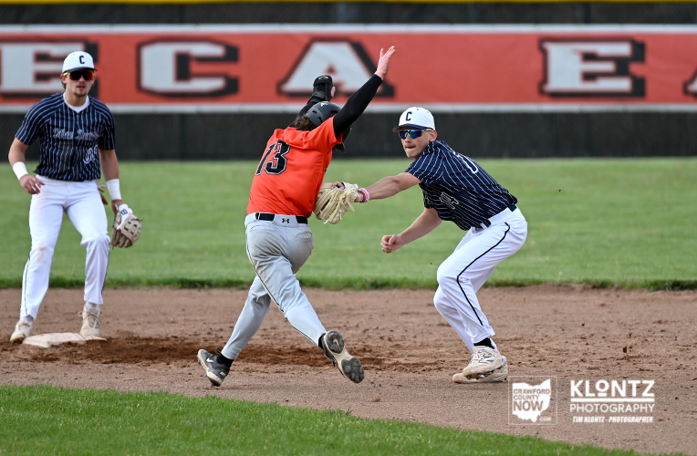 Runner in an orange jersey slides into second base as the shortstop in a striped uniform reaches for a tag; another fielder watches from the left on a dirt infield with green outfield.