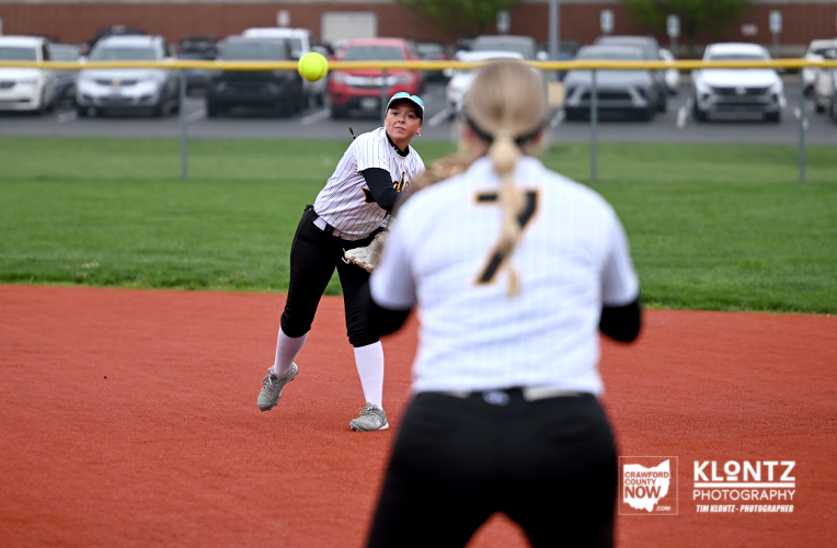 A female softball pitcher throws a ball on a red infield with a blurred teammate wearing number 7 in the foreground and cars in a parking lot behind.