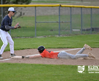 Baseball fielder in a blue-striped uniform watches as a runner in orange slides headfirst into a base on the dirt infield.