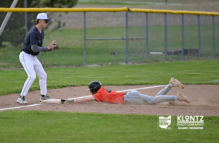 Baseball fielder in a blue-striped uniform watches as a runner in orange slides headfirst into a base on the dirt infield.