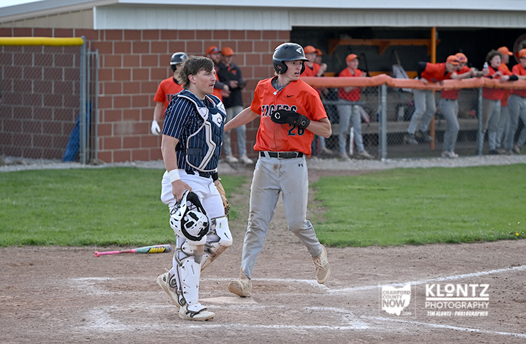 Baseball catcher in blue gear and a batter in an orange uniform greet with a high-five near home plate as teammates watch from the dugout far behind.