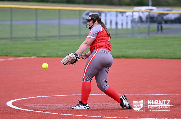 Female softball pitcher in red and gray uniform on the mound, glove ready as a bright ball approaches she prepares to throw or field the pitch.