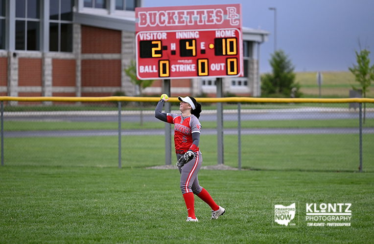 Softball player in red uniform catching a ball on the field with a red scoreboard behind her reading Buckette's and game details.