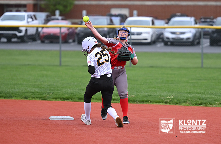 Two young softball players collide near a base on the infield: a white-uniform runner wearing #25 slides into the base while a red-uniform fielder with a glove reaches to tag as the ball flies overhead.