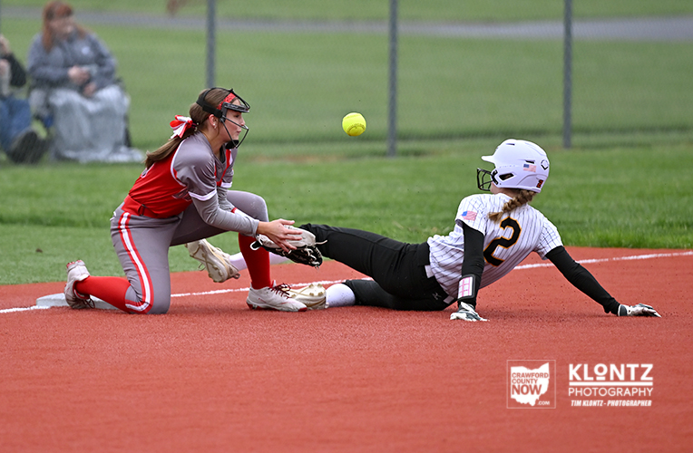Softball play: runner slides into base as catcher reaches for a ground ball during a close play on a dirt infield