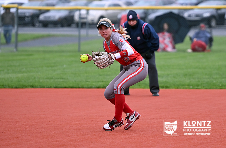 Female softball player in red and gray uniform on the infield, glove outstretched to field a yellow ball.