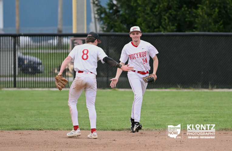 Two Bucyrus baseball players share a high-five on the infield; #14 faces the camera as #8, seen from the back, extends a glove toward him.