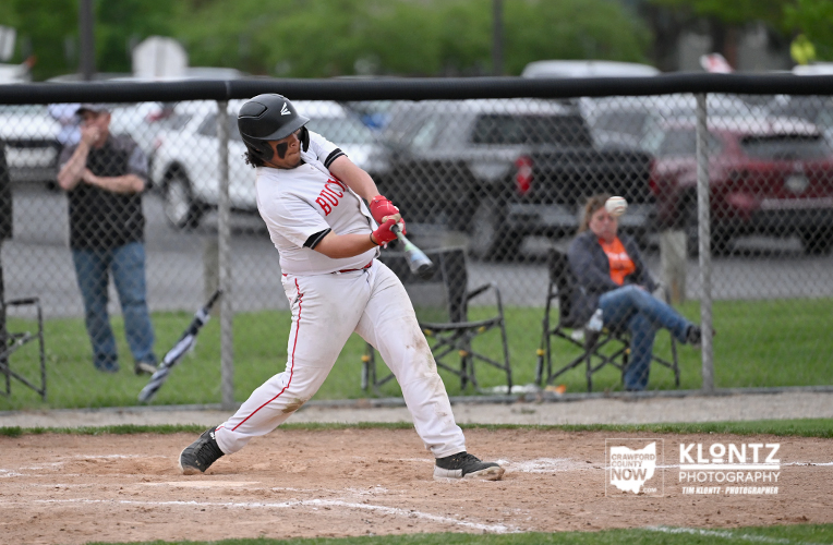 Baseball batter in a white uniform swings at a pitch near home plate with a chain-link fence and spectators in the background.