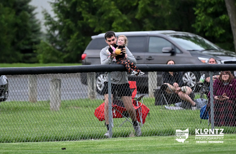 Man carries a smiling toddler over a chain-link fence at a park event, with spectators and a gray SUV in the background.