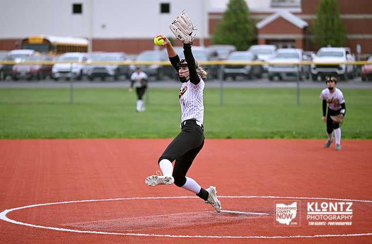 Softball pitcher mid-pitch on the mound, ball in throwing hand and glove raised above head.